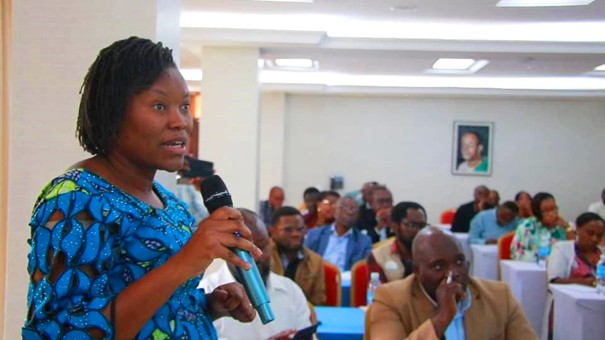 Woman in blue dress holding a microphone, speaking to an audience. 